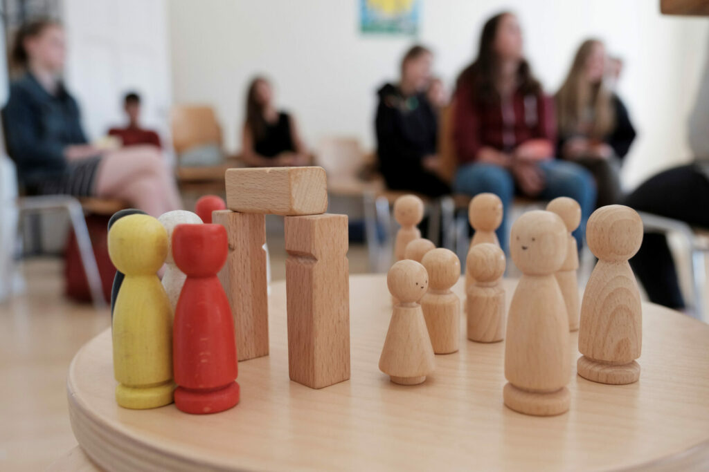 Im Vordergrund eine Gruppe von Holzfiguren im Hintergund Jugendliche in einer Workshopsituation